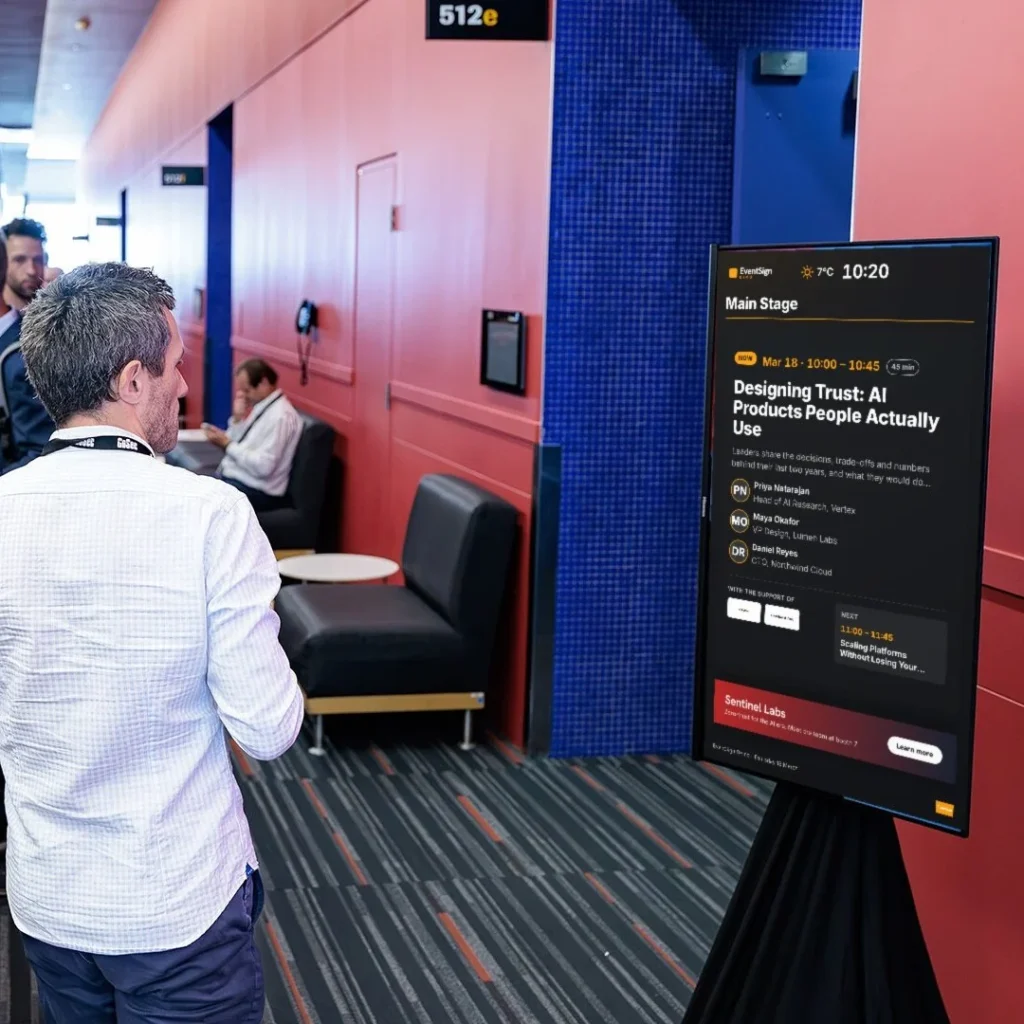 An attendee in a corridor of GoSec 2025 reads a portrait EventSign screen showing the session now on in Main Stage, its speakers and a sponsor banner